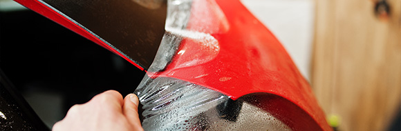 person removing protective film from a red car part showcasing the importance of careful application in car wrapping techniques and the benefits of high-quality materials including seven layers of protection