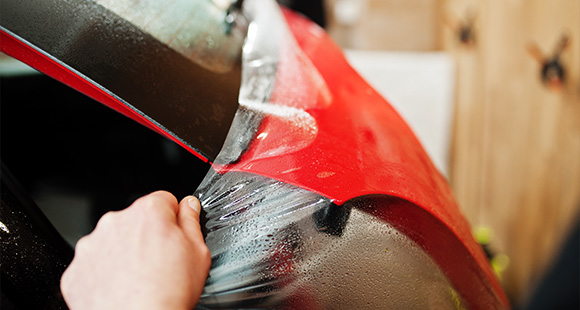 person applying protective film on red car surface showing detailing process for paint protection